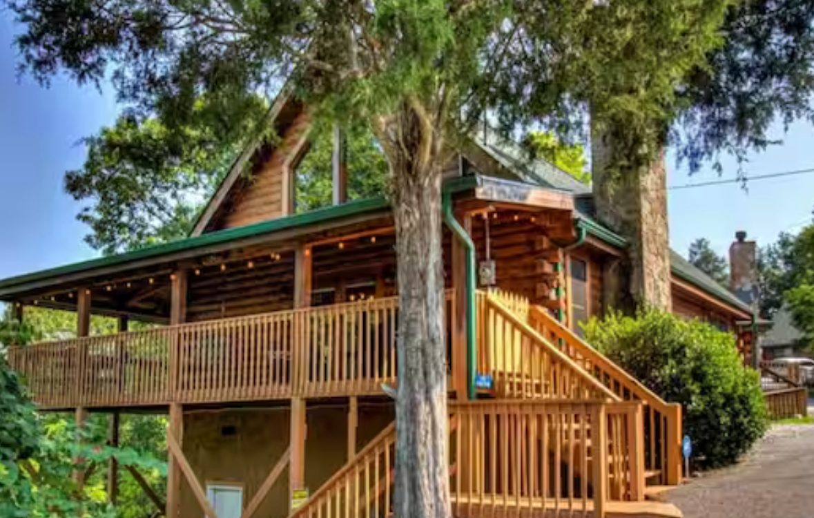 Log cabin exterior with wraparound porch and cedar trees
